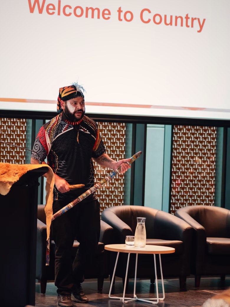 Petiola Wilson delivering a Welcome to Country on stage beside a screen reading Welcome to Country