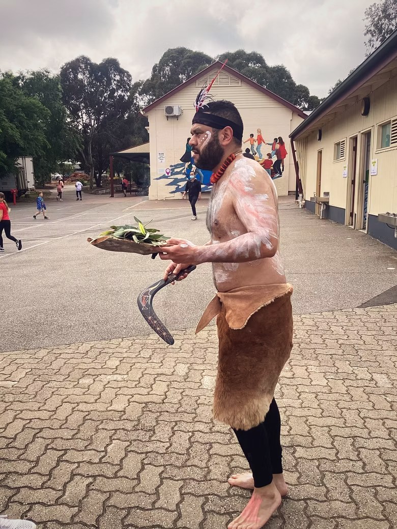 Petiola Wilson in ochre body paint carrying smoking leaves and a boomerang through a school yard